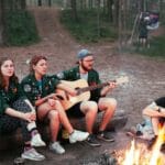 Harcerze siedzą przy ognisku w lesie, jedna osoba gra na gitarze